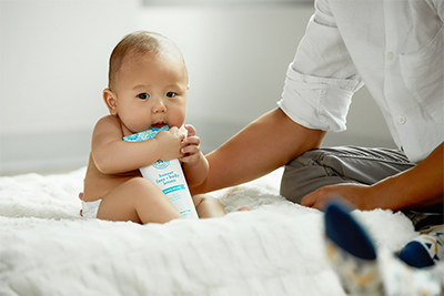 Baby eating a lotion bottle on a white comforter 
