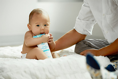 Baby eating a lotion bottle on a white comforter