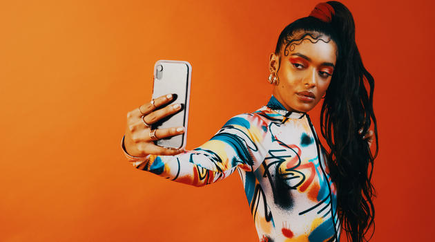 Woman holding phone is outstretched arm, as if she's taking a selfie. She stands in front of a bright orange backdrop.