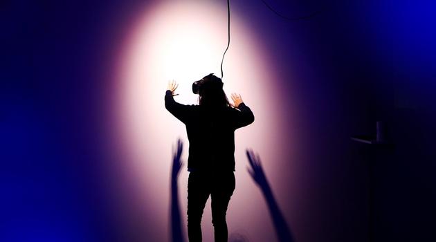 View of person from behind, facing a purple wall. Person is wearing a VR set on their face and has arms outstretched towards the wall.