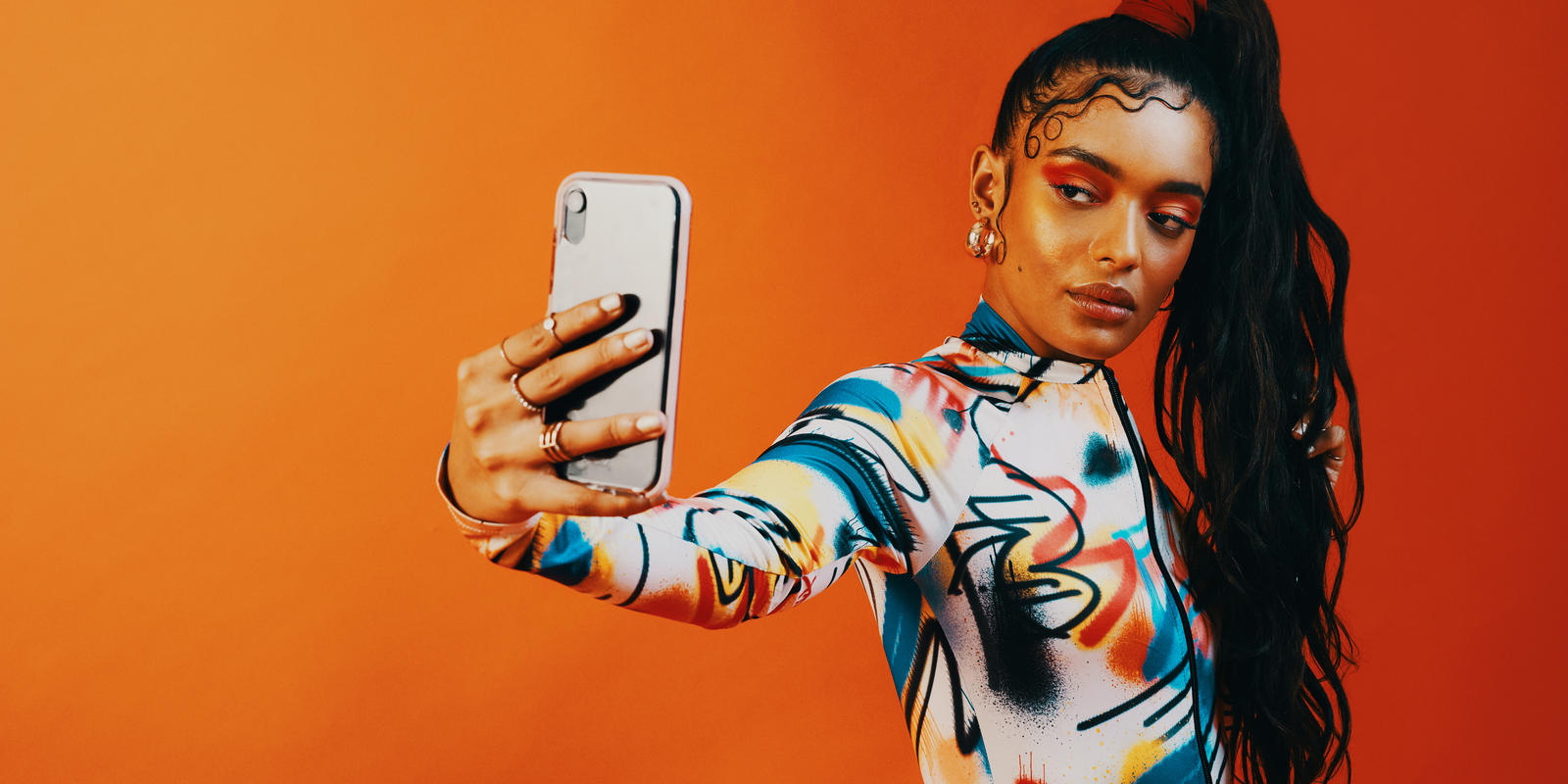 Woman holding phone is outstretched arm, as if she's taking a selfie. She stands in front of a bright orange backdrop.