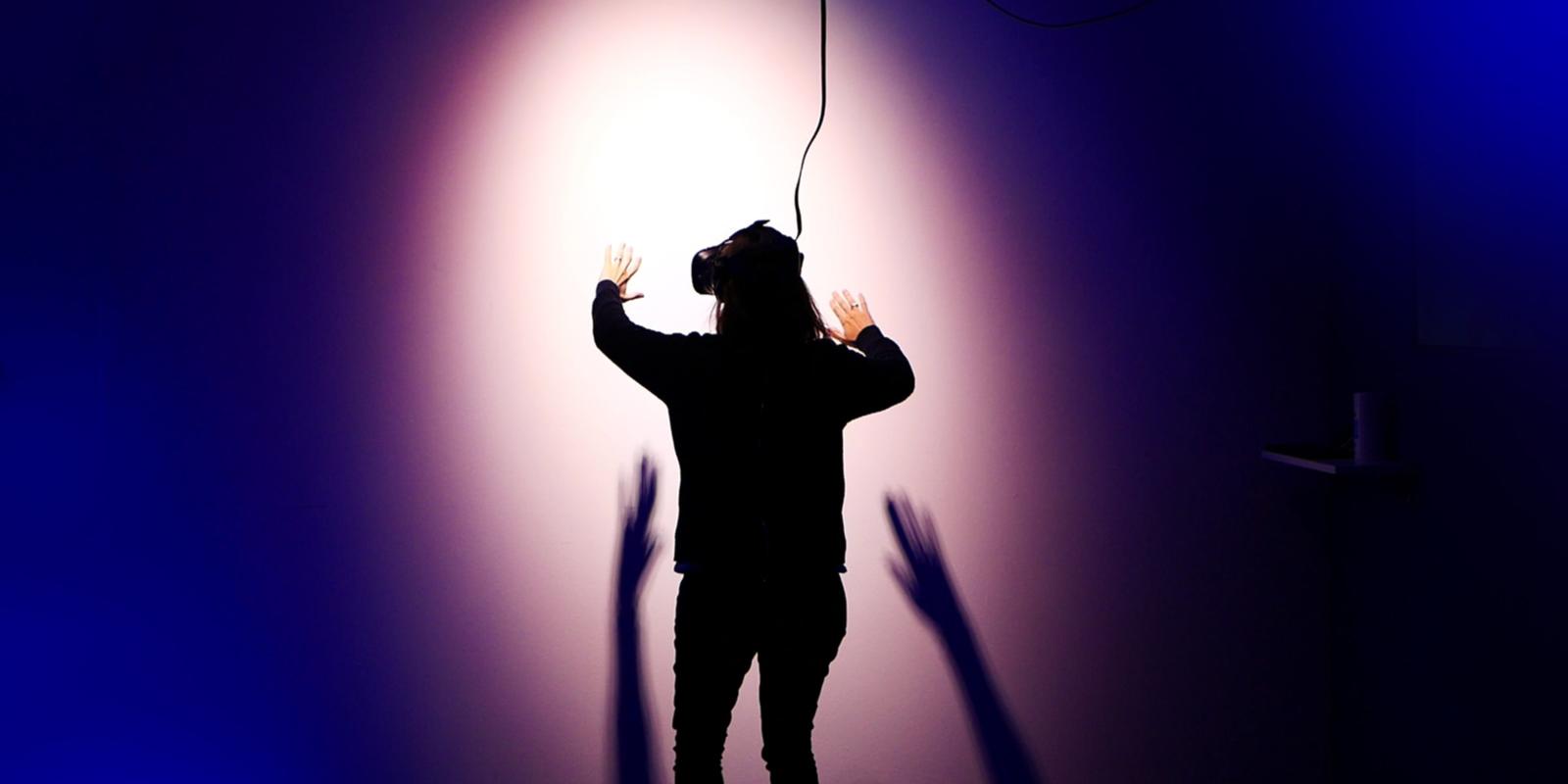 View of person from behind, facing a purple wall. Person is wearing a VR set on their face and has arms outstretched towards the wall.